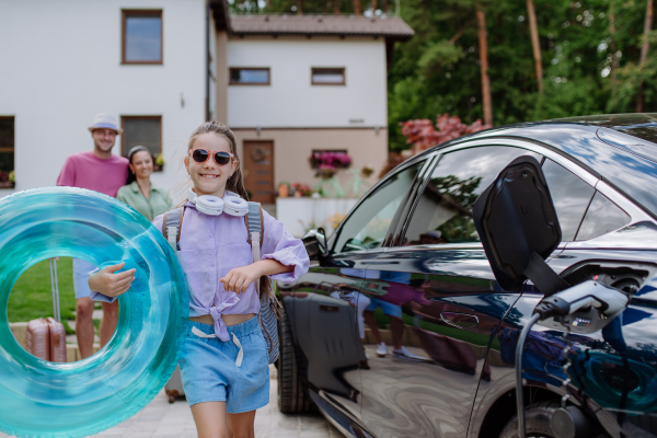 Happy little girl posing with swimming wheel, preparing for holiday while the electric car charging.