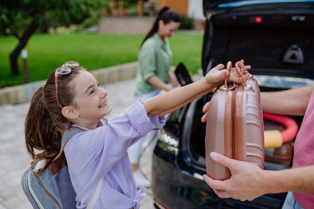 Happy mother holding power supply cable and charging their electric car, rest of family putting suitcases in a car trunk, preparating for holidays.