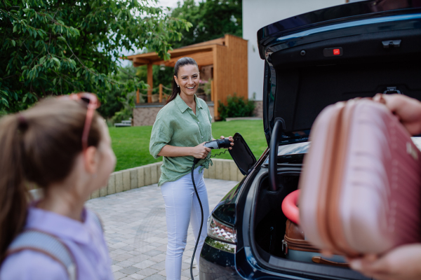 Happy mother holding power supply cable and charging their electric car, rest of family putting suitcases in a car trunk, preparating for holidays.