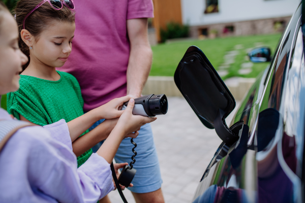 Close-up of father holding power supply cable with his little daughters and charging electric car.