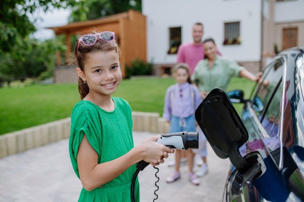 Little girl holding power supply cable and charging electric car, rest of family waiting in background.