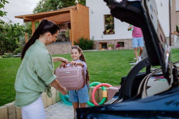 Happy family preparing for a holiday, putting suitcases in a car trunk, while their electric car charging.