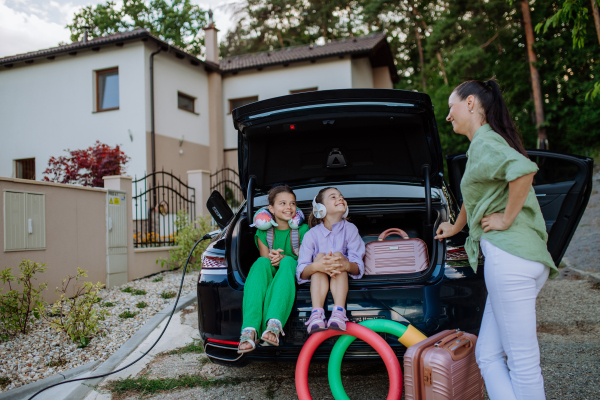 Happy sisters with their mother sitting in a car trunk and waiting for electric car charging.