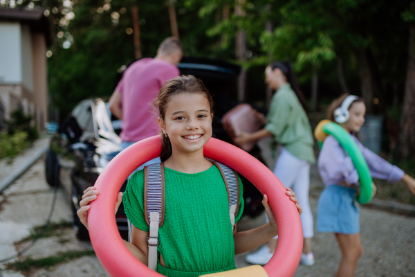 Happy family preparing for a holiday, putting suitcases in a car trunk, while their electric car charging.Little daughter posig with headphones and swimming wheel.