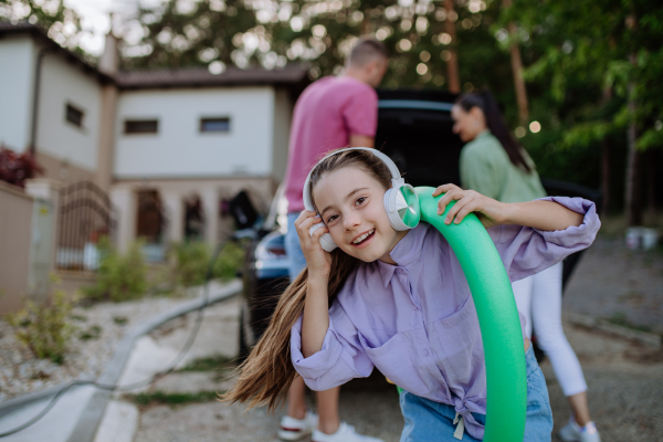 Happy family preparing for a holiday, putting suitcases in a car trunk, while their electric car charging.Little daughter posig with headphones and swimming wheel.