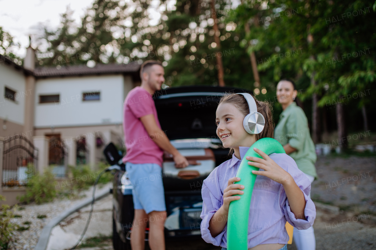 Happy family preparing for a holiday, putting suitcases in a car trunk, while their electric car charging.