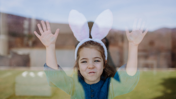 Portrait of little girl with bunny ears standing behind a window.