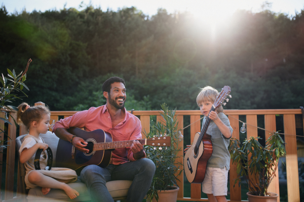 A young African american man enjoys playing the guitar with little children accompaining him with instruments on patio in garden in summer.