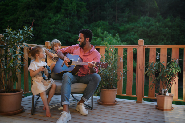 A young African american man enjoys playing the guitar with little children accompaining him with instruments on patio in garden in summer.