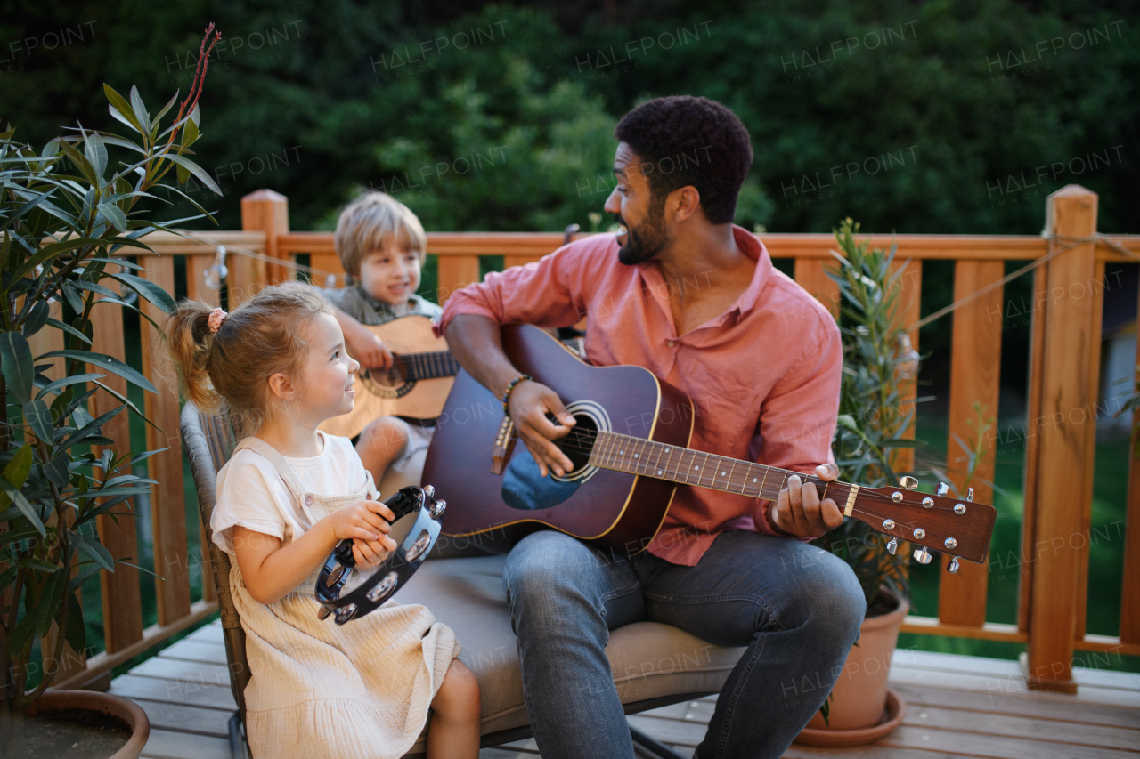 A young African american man enjoys playing the guitar with little children accompaining him with instruments on patio in garden in summer.