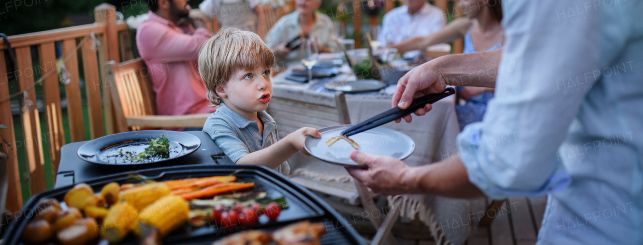 A father putting grilled meat and vegetable on plate to his son during family summer garden party.