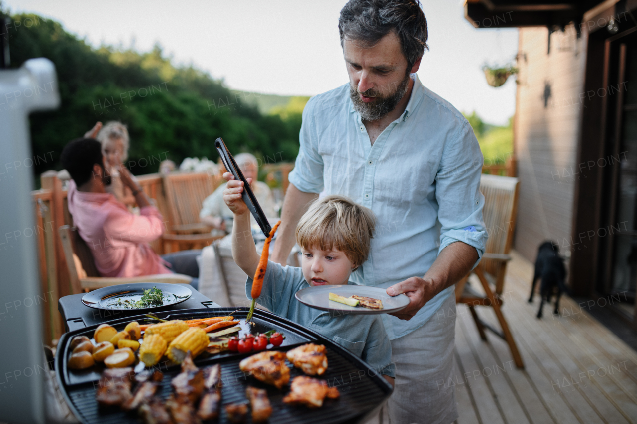 An father with little son grilling ribs and vegetable on grill during family summer garden party.