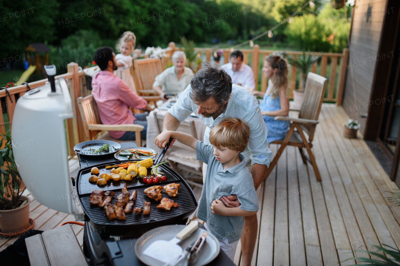 An father with little son grilling ribs and vegetable on grill during family summer garden party.