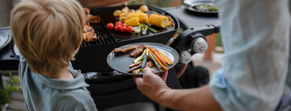 A father putting grilled meat and vegetable on plate to his son during family summer garden party.