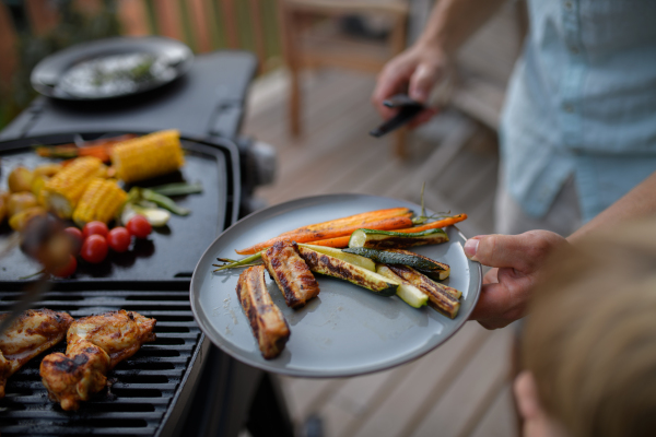 A father putting grilled meat and vegetable on plate to his son during family summer garden party.