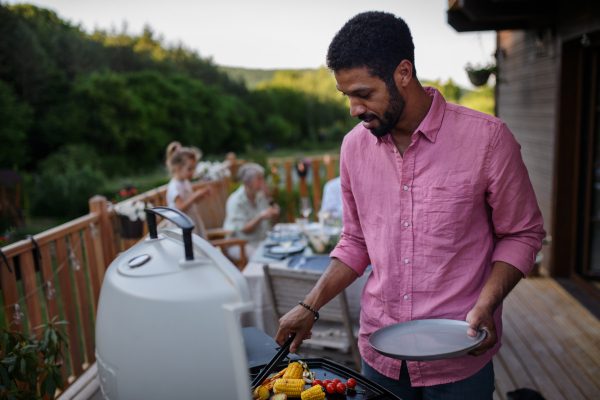 A father grilling meat and vegetable on grill during family summer garden party.