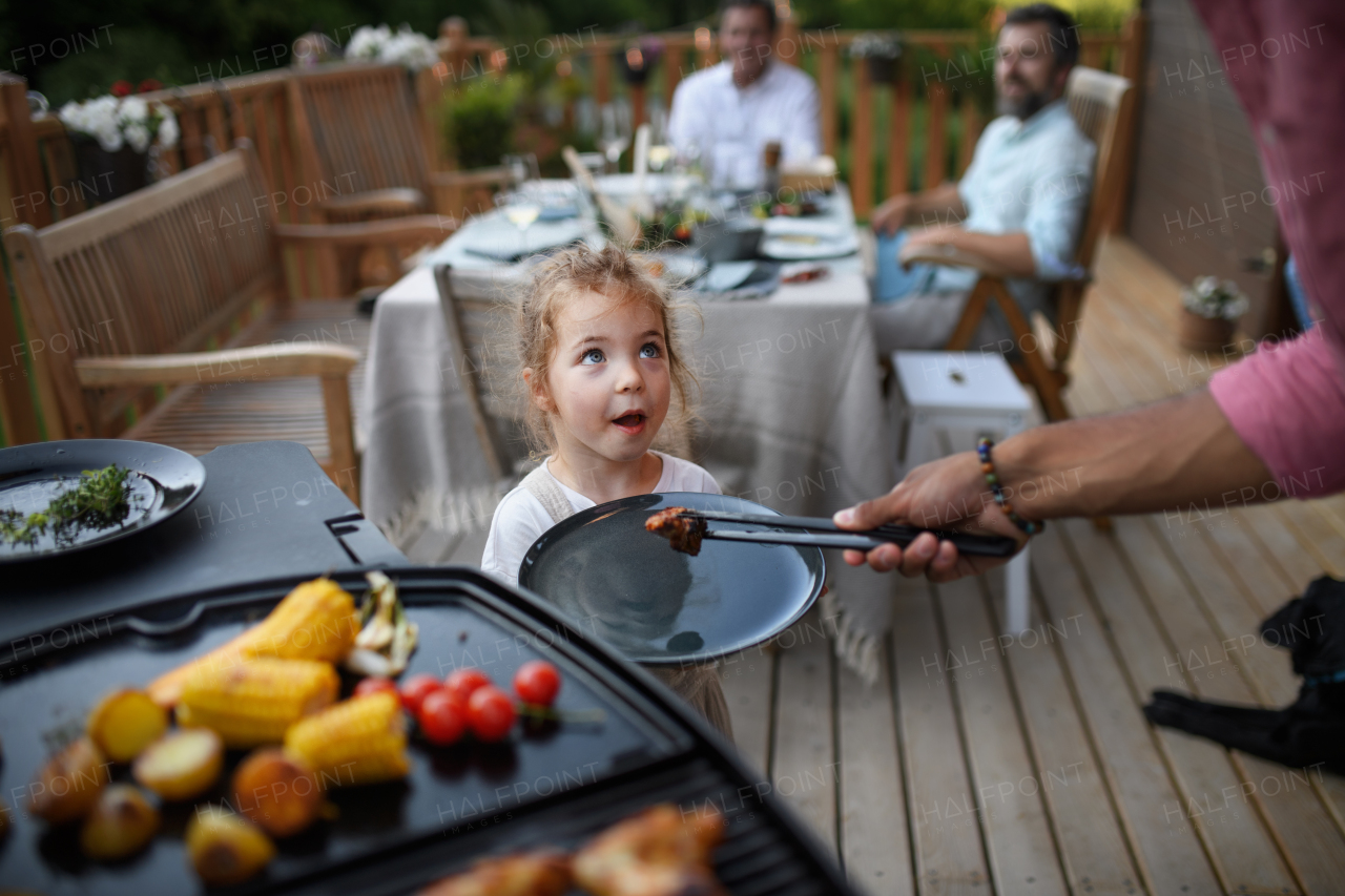 A father putting grilled meat and vegetable on plate to his daughter during family summer garden party.