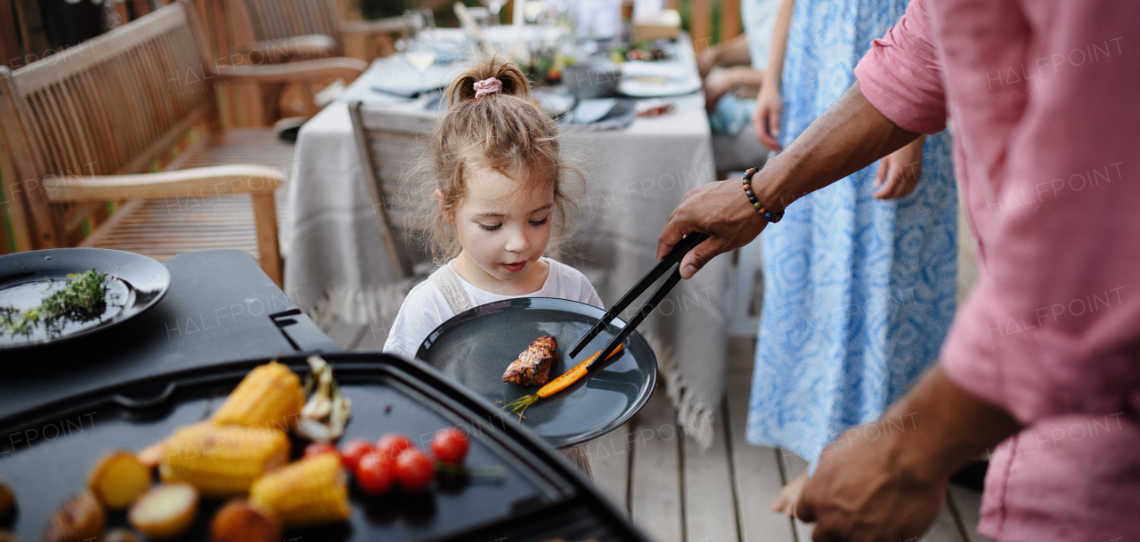 A father putting grilled meat and vegetable on plate to his daughter during family summer garden party.