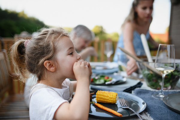 A family eating at barbecue party dinner on patio, little girl eating roasted corn and enjoying it.