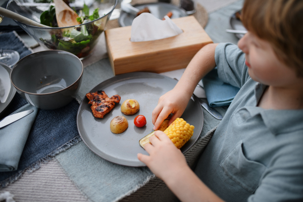 A little boy eating roasted corn and vegetables at family barbecue party in garden.