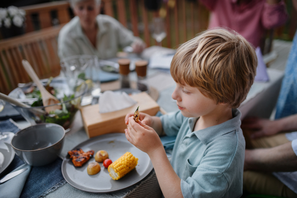 A little boy eating roasted corn and vegetables at family barbecue party in garden.