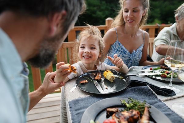 A family eating at barbecue party dinner on patio, little girl with parents sitting at table and enjoying food.