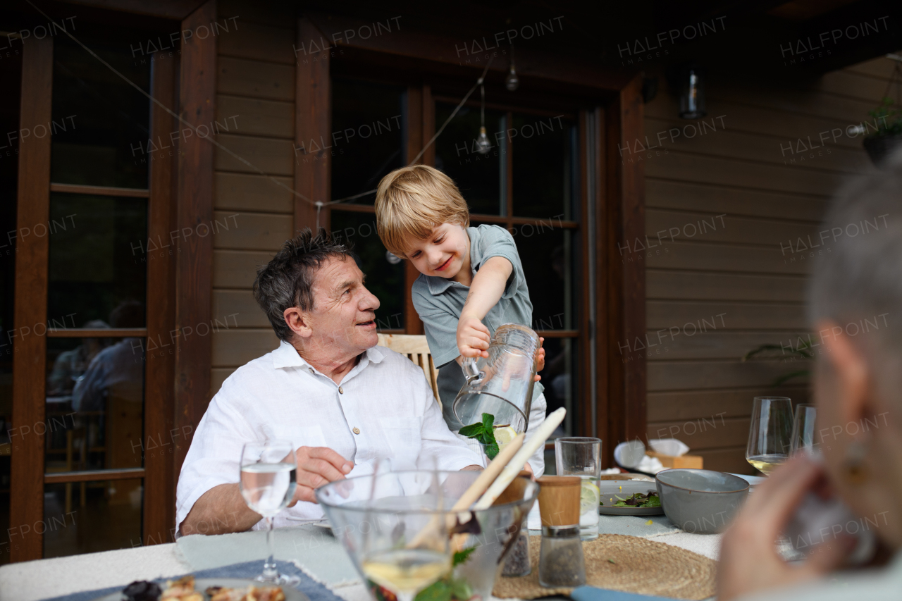 A little boy pouring lemonade to his grandfather during family celebration outside on patio.