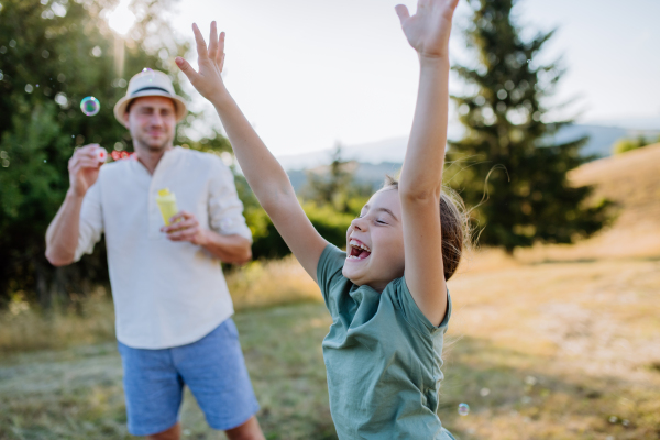 A young father and his little daughter having fun while blowing soap bubbles on a summer day in nature.