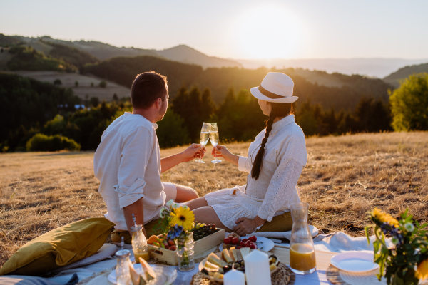 An attractive couple in love enjoying picnic and drinking wine on the hill at sunset.