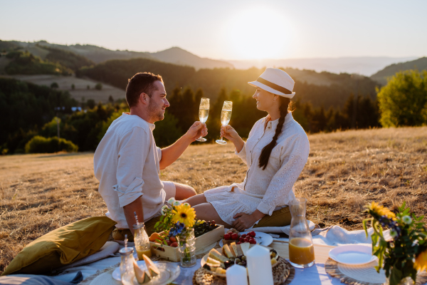 An attractive couple in love enjoying picnic and drinking wine on the hill at sunset.