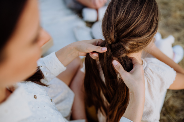 Close-up of mother making hairstyle to her daughter during family picnic in the nature.