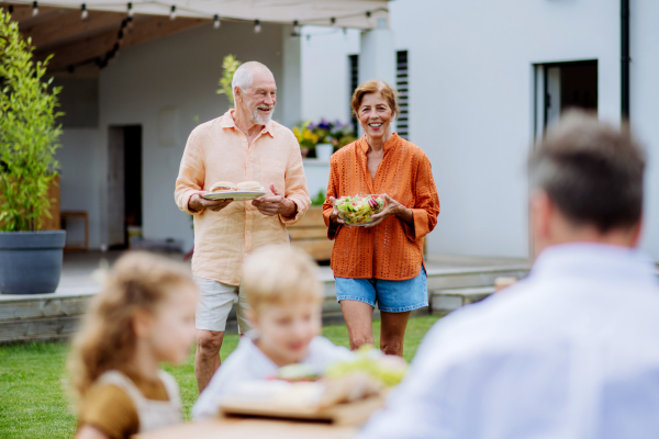 Happy senior couple bringing a salad and burgers at multi generation garden party in summer.