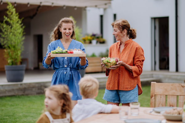 Happy women bringing a salad and burgers at multi generation garden party in summer.