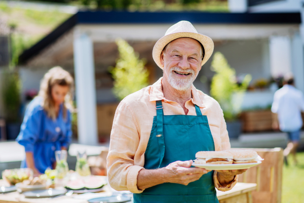 A happy senior man serving burgers at multi generation garden party in summer.