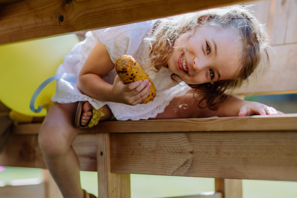 A cute little girl eating grilled corn and looking under the table at camera during garden party.