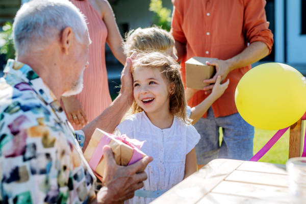 A happy little girl giving birthday present to her senior grandfather at generation family birthday party in summer garden