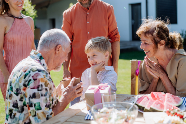 A happy little boy giving birthday present to his senior grandfather at generation family birthday party in summer garden