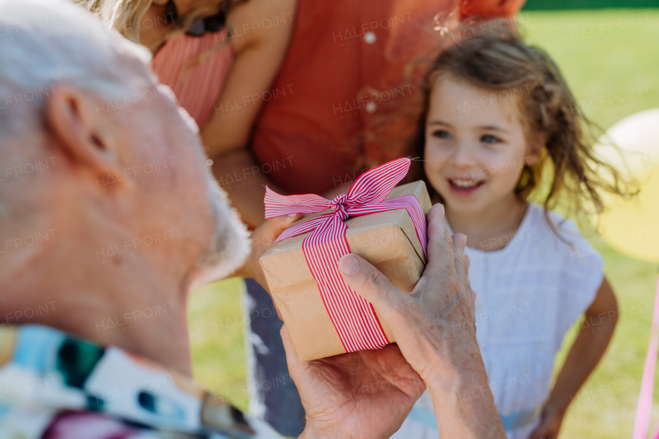 A happy little girl giving birthday present to her senior grandfather at multigenerational family birthday party in summer garden.