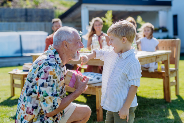 A happy little boy giving birthday present to his senior grandfather at generation family birthday party in summer garden