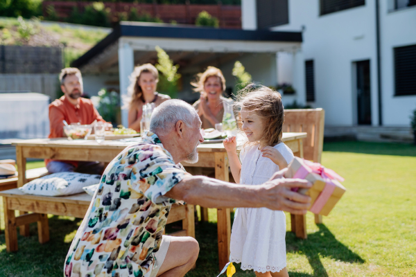 A happy little girl giving birthday present to her senior grandfather at generation family birthday party in summer garden.