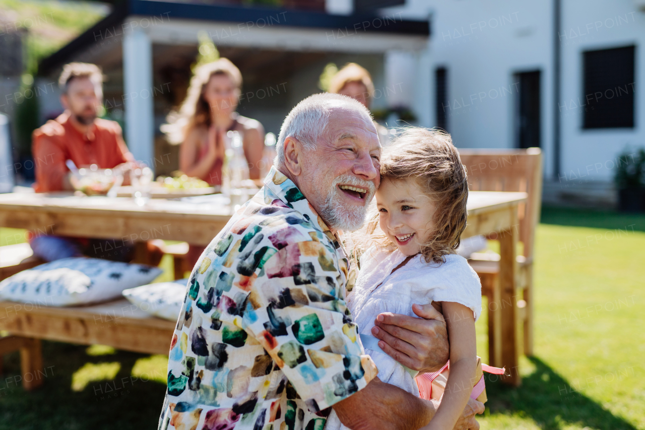 A happy little girl giving birthday present to her senior grandfather at generation family birthday party in summer garden