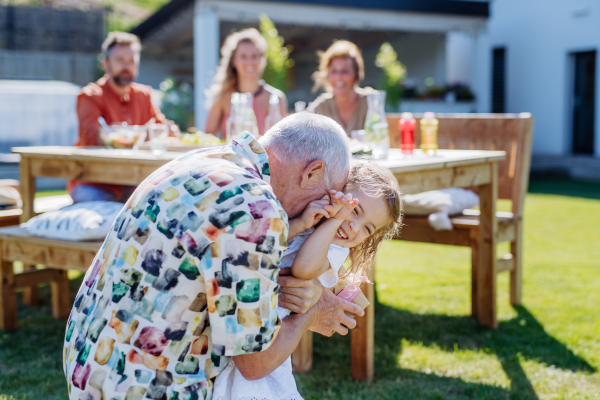 A happy little girl giving birthday present to her senior grandfather at generation family birthday party in summer garden