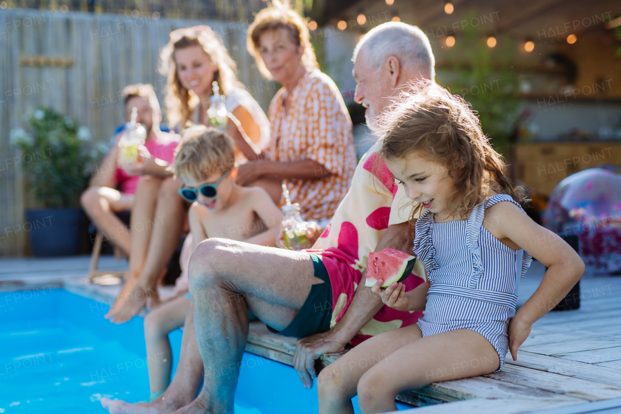 A multi generation family enjoy watermelon and drinks near backyard pool.