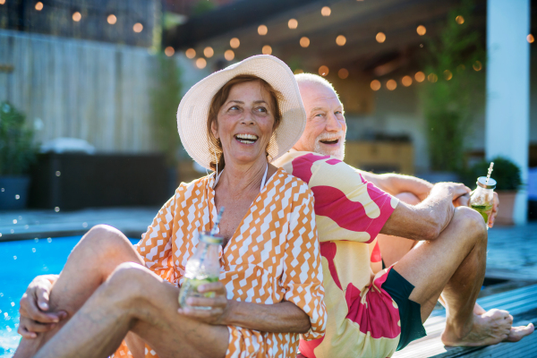 A happy senior couple enjoying drinks when relaxing and sitting by swimming pool in summer.