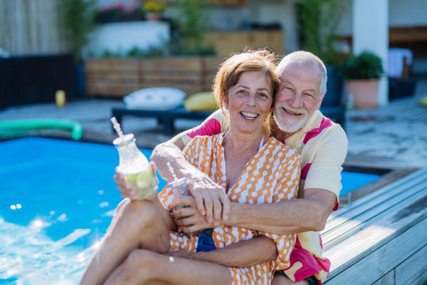 A happy senior couple enjoying drinks when relaxing and sitting by swimming pool in summer, looking at camera.