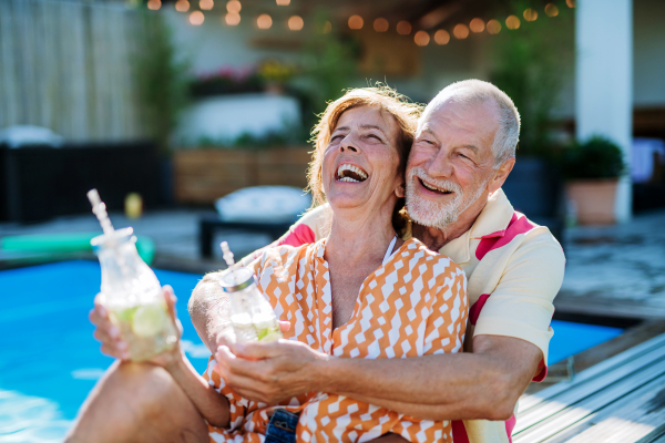 A happy senior couple enjoying drinks when relaxing and sitting by swimming pool in summer.