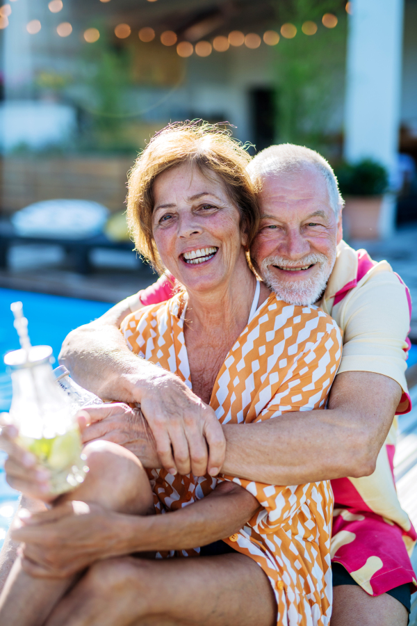 A happy senior couple enjoying drinks when relaxing and sitting by swimming pool in summer.