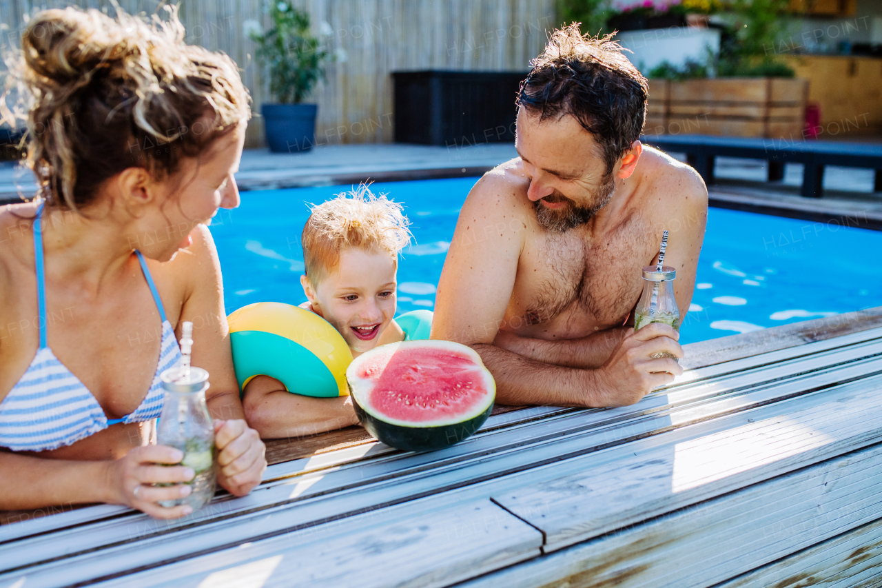 A happy little boy with his parents in backyard swimming pool enjoying refreshments, drinks and watermelon.