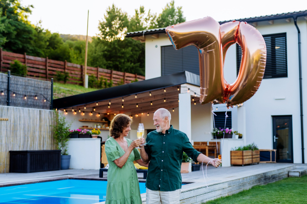 A man with number 70 balloons with his wife celebrating birthday and toasting with wine near backyard pool.