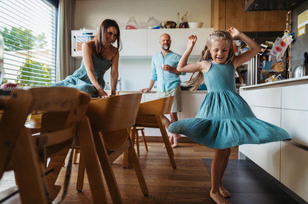 Little girl dancing in the kitchen, while parents watching her. Cheerful family having fun together in kitchen on sunny morning. Concept of family bonding.